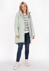 Giacca impermeabile verde chiaro con cappuccio, dotata di bottoni a scatto, due tasche laterali e vestibilità comoda, indossata sopra una maglietta a righe e jeans.