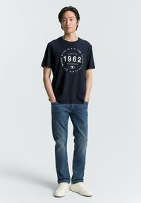 Camiseta de algodón azul marino con un gráfico circular blanco que dice '1962' y 'Casual Classics'. Combinada con unos vaqueros de mezclilla azul claro y zapatillas blancas.