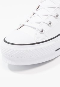Białe płócienne sneakersy z gumową podeszwą, okrągłym noskiem, płaskim sznurowadłem i teksturowanym poziomym paskiem na krawędzi podeszwy. Czarny detal wykończeniowy.