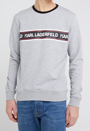 Sweatshirt gris clair avec un col rond noir, arborant une bande horizontale noire et blanche avec le texte "KARL LAGERFELD" en gras. Matière en mélange de coton.