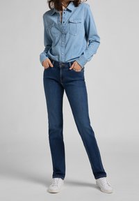 Persona che indossa una camicia di jeans azzurra chiara infilata in jeans dritti blu scuro con sneakers bianche, mani nelle tasche contro uno sfondo semplice.