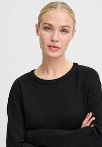 Pull noir à col rond, avec des poignets côtelés et une coupe décontractée, fabriqué en tissu doux. Le modèle a les bras croisés, mettant en valeur le vêtement.