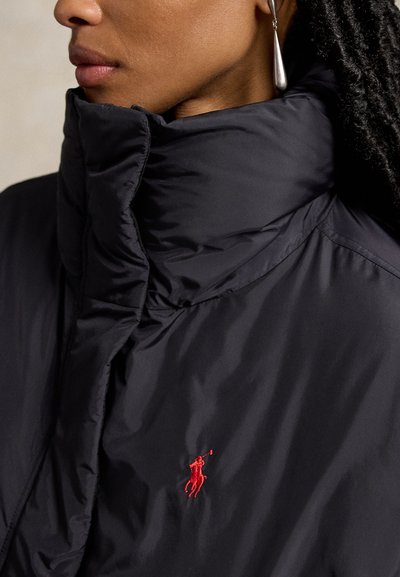 Gros plan d'une personne portant une doudoune noire avec un col montant et un petit logo rouge brodé de joueur de polo sur la poitrine.