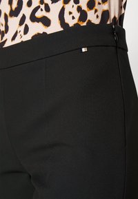 Czarne spodnie o gładkiej fakturze, z subtelnym połyskiem, dopasowanym krojem i małym ozdobnym akcentem w pobliżu talii.