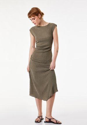 Vrouw met kort rood haar, gekleed in een mouwloze olijfgroene midi-jurk en zwarte sandalen, staand en naar beneden zijwaarts kijkend.