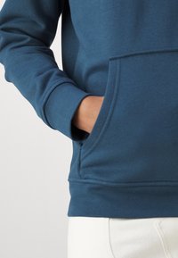 Zils vilnas džemperis ar kangaroo kabatu un ribotām piedurknēm, demonstrējot mīkstu tekstūru un brīvu piegriezumu. Balti bikses daļēji redzamas.