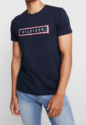 Dunkelblaues T-Shirt aus Baumwolle mit einem rot umrandeten rechteckigen Logo, das "HILFIGER" in weißer Schrift auf der Brust zentriert zeigt. Kurze Ärmel.