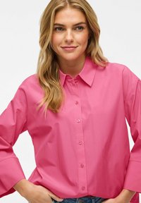 Camicia button-up rosa brillante realizzata in tessuto liscio con colletto a punta e maniche ampie, caratterizzata da una fila di bottoni sul davanti.
