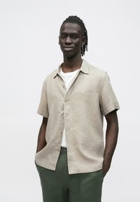 Beiges Leinenhemd mit kurzen Ärmeln und durchgehender Knopfleiste, kombiniert mit einem weißen T-Shirt und dunkelgrünen Hosen. Einfaches Kragen-Design.