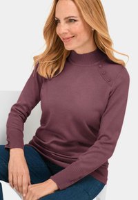 Pull en tricot bordeaux avec un col montant, des poignets côtelés et un détail de trois boutons à l'épaule, porté avec un jean bleu.