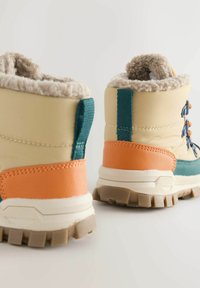 Bottes imperméables beiges avec des accents turquoise et orange, doublure en polaire et semelle en caoutchouc texturée ; elles disposent d'un lacet robuste et d'un talon renforcé.