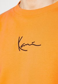 Orange bomullströja med en svart broderad signaturlogotyp på bröstet. Ribbad halsringning och slät textur.