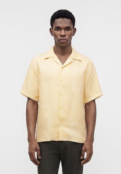 Jeune homme portant une chemise jaune clair à manches courtes boutonnée et un pantalon foncé, debout devant un fond clair uni.