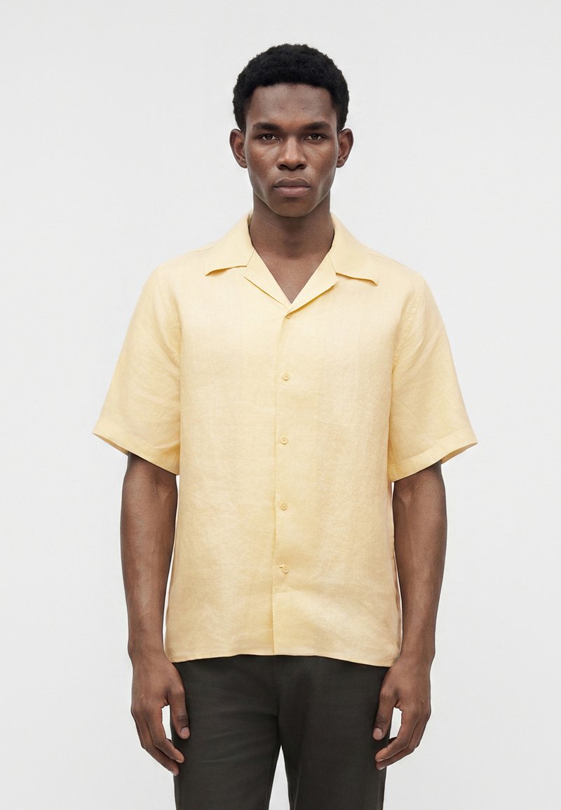 Jeune homme portant une chemise jaune clair à manches courtes boutonnée et un pantalon foncé, debout devant un fond clair uni.