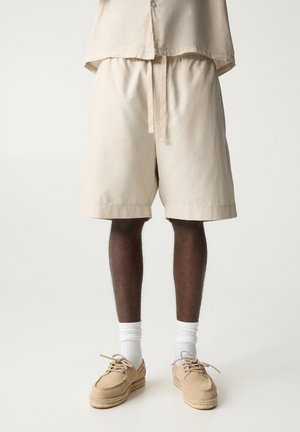 Beige cotton shorts with an elastic waistband and drawstring. Paired with beige shoes and white socks. Plain, knee-length design.