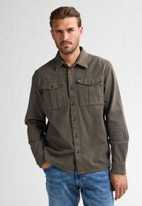 Olive green button-up shirt with two chest pockets, brown buttons, and a soft texture. Worn with light blue jeans, displayed standing.