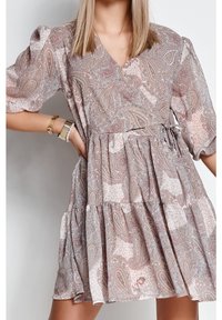 Kobieta w luźnej sukience do kolan z bufiastymi rękawami w różowo-beżowy wzór paisley, uzupełnionej bransoletkami.