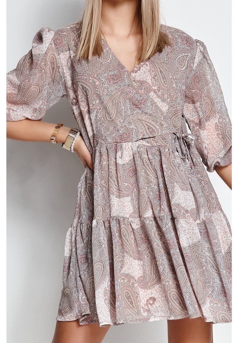 Kobieta w luźnej sukience do kolan z bufiastymi rękawami w różowo-beżowy wzór paisley, uzupełnionej bransoletkami.