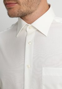 Mann trägt ein makelloses weißes Hemd mit spitzem Kragen, zugeknöpft, zeigt das untere Gesicht und leichtes Gesichtshaar.