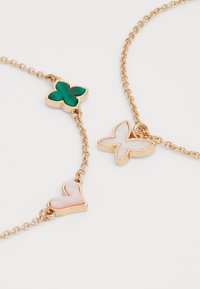 Pulsera de cadena en tono dorado que presenta tres charms: una mariposa verde, un corazón rosa y una mariposa blanca, cada uno contornado en dorado.