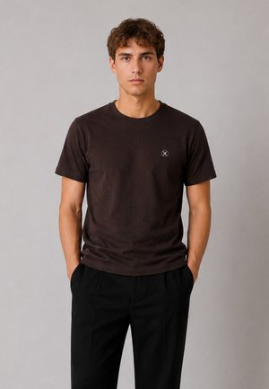 Jeune homme debout, les mains dans les poches, portant un t-shirt marron foncé uni et un pantalon noir sur un fond gris clair.