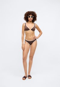 Brown bikini with white trim, featuring a triangular top with a knot detail. Worn with dark slides and white sunglasses, against a neutral background.