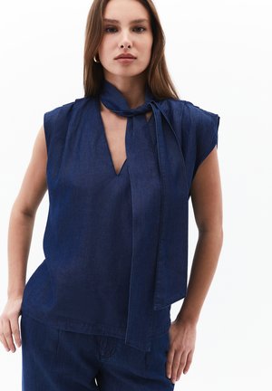 Top denim gelap tanpa lengan dengan leher V yang dalam dan detail kerah yang diikat. Tekstur halus, potongan santai, dan celah di sisi hem.