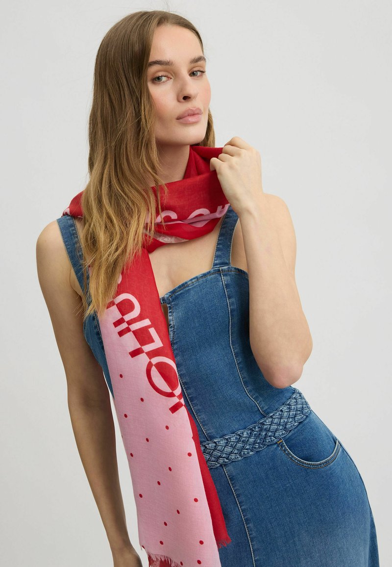 Rode en roze sjaal met een stippenpatroon en vetgedrukte tekst, gemaakt van een zachte stof, om de nek van het model gedrapeerd, gecombineerd met een denim jurk.
