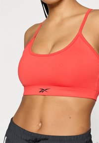 Κοραλλί αθλητικό σουτιέν με λεπτές τιράντες, στρογγυλή λαιμόκοψη και λογότυπο Reebok σε μαύρο στη λωρίδα. Λείο, ελαστικό ύφασμα για άνεση.