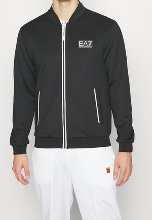 Mand iført en sort EA7 Emporio Armani lynlåstrøje med hvide kanter og hvide bukser med et lille rødt og gult logo.