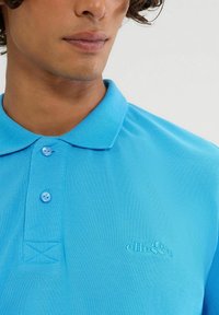 Himmelblaues Poloshirt aus strukturiertem Stoff, mit Kragen, drei Knöpfen und einem geprägten Logo auf der Brust.