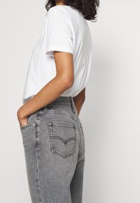 Jeans grises con un diseño de pierna recta, hechos de denim, que presentan una forma clásica de bolsillo trasero y una pequeña etiqueta de logo roja. Combinados con una camiseta blanca.
