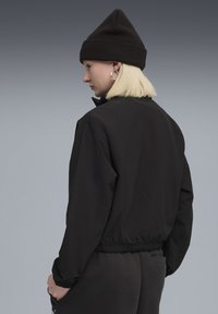 Person med kort blond hår iført en sort beanie, sort jakke og mørke bukser, stående sidelæns mod en grå baggrund.