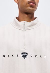 Άνδρας που φοράει λευκό πουλόβερ Nike Golf με φερμουάρ μέχρι το στήθος, με μαύρο κεντημένο λογότυπο και κείμενο στο στήθος.