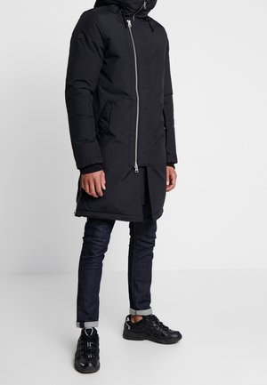 Personne portant un long manteau noir matelassé avec une fermeture éclair à l'avant, un jean foncé retroussé et des baskets noires, debout sur un sol blanc uni.