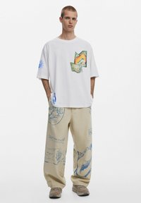 Balta medvilninė marškinė su spalvingomis grafikos detalėmis priekyje ir ant rankovių, derinamos su béžinėmis kelnėmis, išryškinančiomis mėlynus raštus ir akcentus.