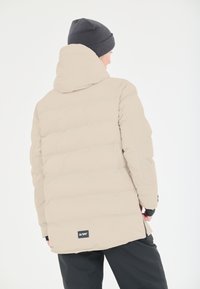 Ljust beige puffjacka med huva, med horisontella quiltade sektioner och en logotyp-patch vid nederkanten. Mörk mössa bärs på huvudet.