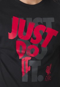 T-shirt noir avec texte superposé en gras « JUST DO IT. » en rouge et gris, avec le logo oiseau du Liverpool FC en rouge en bas à droite.