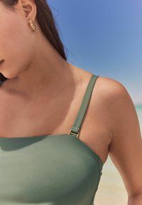 Grüner Badeanzug mit strukturiertem Design, schmalen Trägern und goldfarbener Verschlussdetail. Glatte Textur vor einem sanften Strandhintergrund.