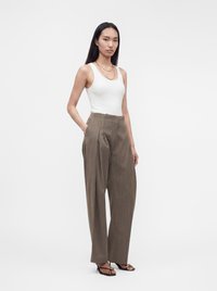 Femme debout, les mains dans les poches, portant un haut blanc sans manches, un pantalon plissé marron ample, des sandales noires et des colliers dorés superposés.