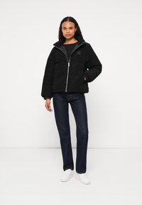 Czarna pikowana kurtka puchowa z wysokim kołnierzem, srebrnym zamkiem i prążkowanymi mankietami, zestawiona z ciemnymi dżinsami i białymi trampkami.