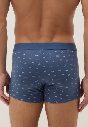 Männlicher Torso von hinten, der blaue Boxershorts mit kleinen weißen Wellenmustern vor einem neutralen Hintergrund trägt.