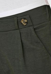 Pantalones verde oscuro con cierre de botón frontal, detalle de pliegues, bolsillo lateral y una camisa blanca metida en la cintura.