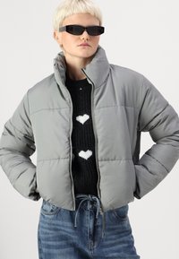 Giubbotto imbottito grigio con colletto alto, chiusura a zip, design trapuntato. Abbinato a un maglione nero con motivi a cuore bianchi e jeans in denim.