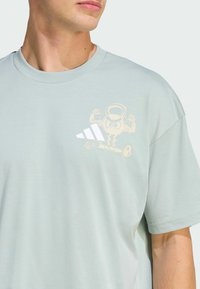 Light mint green cotton t-shirt featuring a graphic of a cartoon character lifting weights and three white stripes on the chest.