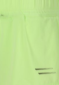 Neongröna sportshorts med en slät textur, elastisk midja och reflekterande svarta detaljer med ett mönstrat mönster på sidan.