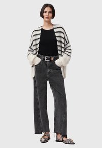 Gestreepte, oversized cardigan in crème en zwart; zwarte t-shirt; hoge taille, brede zwarte jeans; zwart-witte schoenen met gouden accenten.