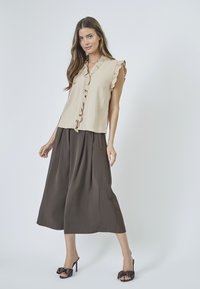 Beige ärmelloses Top mit rüschengeschnittenen Kanten, kombiniert mit braunen weiten Hosen. Die Schuhe sind schwarze Sandalen mit Schleifenakzenten.