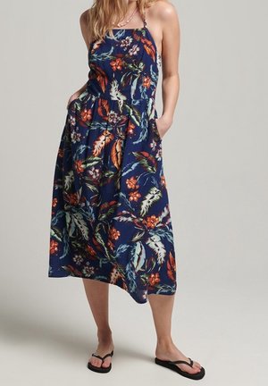 Vrouw die een marineblauwe bloemenmouwloze midi-jurk met zakken en zwarte teenslippers draagt, staand tegen een effen achtergrond.