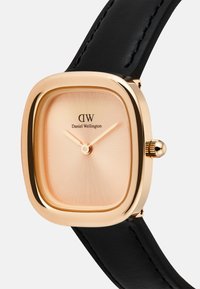 Montre carrée avec un boîtier en métal doré rose, un cadran beige en éclat de soleil, un bracelet en cuir noir, et un détail de couronne sur le côté. Nom de la marque : Daniel Wellington.
